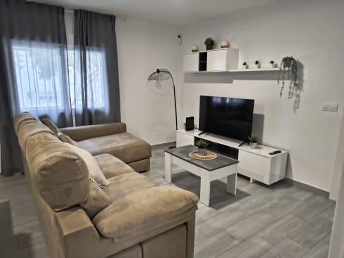Moderno salón de apartamento en Puerto Santa María con sofá beige, televisor, estanterías blancas y vistas al jardín