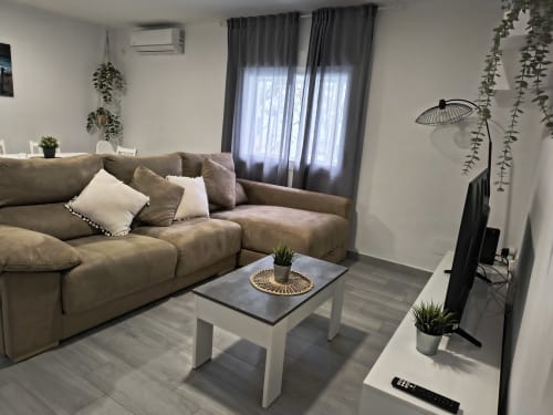 Sala moderna con sofá seccional beige, mesa de café blanca, cortinas grises, plantas en macetas y televisor de pantalla plana