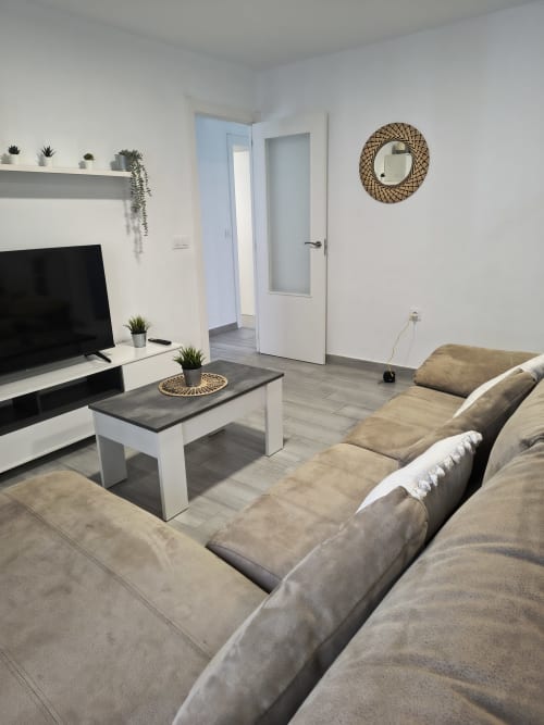 Moderno salón con sofá beige, TV, plantas decorativas y espejo tejido en Wehost Costaluz Puerto Santa María