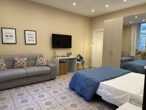 Camera d'hotel Encanto a Napoli con letto, divano grigio, TV, guardaroba con specchio e quadri motivazionali