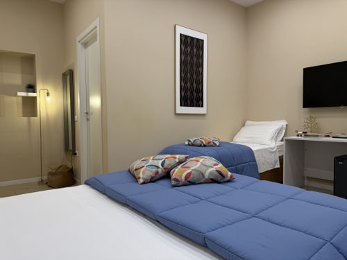 Camera moderna dell'hotel Encanto a Napoli con due letti, coperte blu, cuscini decorati, quadri e TV a schermo piatto