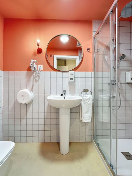 Modern bathroom at Lemon Hostel Altea with orange walls, round mirror, pedestal sink, white tiles, and glass shower enclosure
