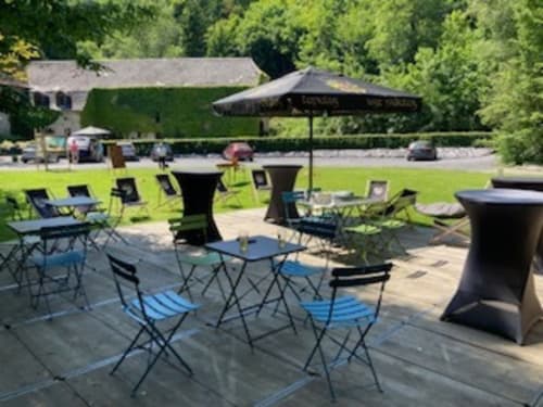 Terrasse extérieure de l'hôtel Le Moulin Des Ramiers à Crupet avec chaises bleues, tables, parasol noir et pelouse verte.
