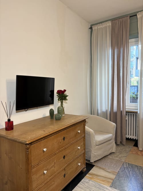 Camera moderna all'Hotel Le Arche Verona con comò in legno, TV a schermo piatto, fiori in vaso, poltrona color crema e vista sulla città