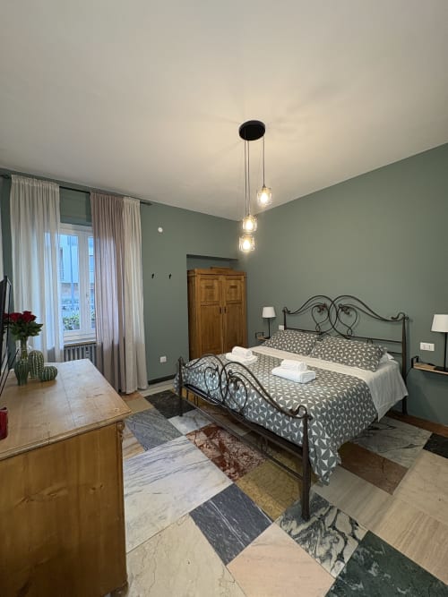 Camera elegante all'hotel Le Arche di Verona con pareti verdi, letto in ferro battuto, luci a sospensione e vista sulla città dalle finestre.
