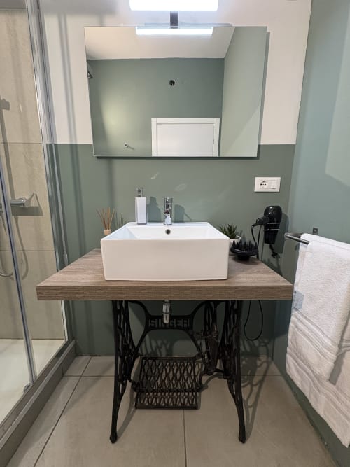 Bagno moderno all'hotel Le Arche di Verona con lavabo bianco a bacinella, pareti verde salvia e mobiletto vintage