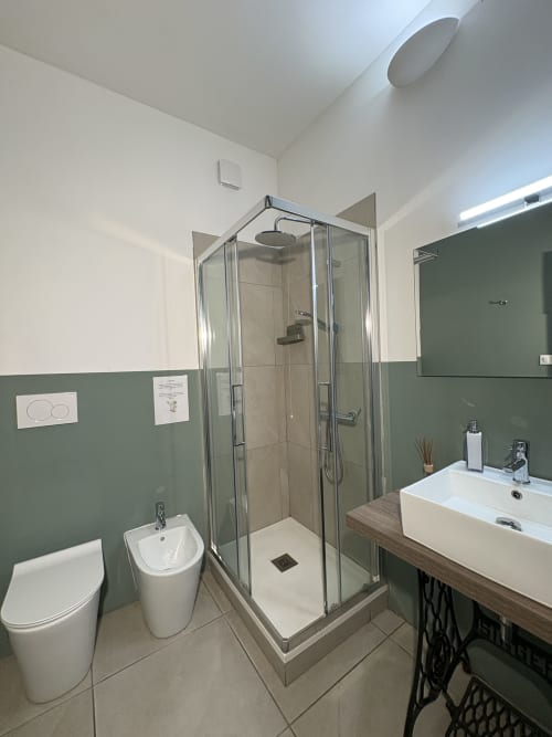 Bagno moderno dell'hotel Le Arche a Verona con box doccia in vetro, lavabo e wc bianchi, rivestimento murale verde