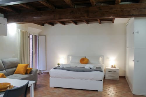 Modern bedroom in Ferrara hotel with white bed, gray sofa, wooden ceiling beams, and terracotta floors