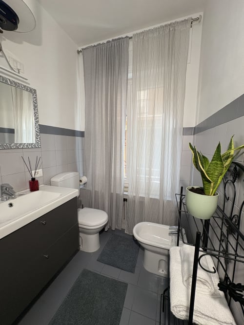 Bagno moderno dell'hotel Le Arche a Verona con pavimento in piastrelle scure, sanitari bianchi, tende grigie e una pianta di sansevieria