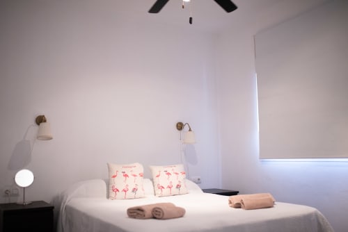 Moderna habitación de apartamento en Mérida con cama blanca, cojines flamenco, ventilador de techo y apliques de pared