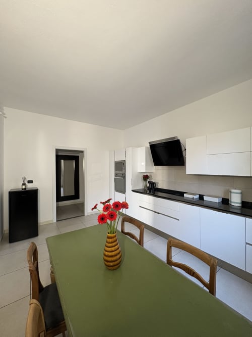 Moderna zona cucina-pranzo dell'appartamento Le Arche Verona con tavolo verde, fiori rossi e mobili bianchi