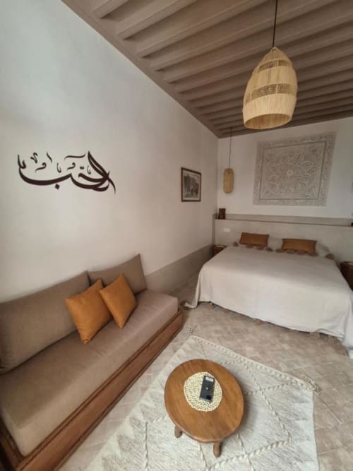 Chambre Hana Suite au Riad Casa Angelo à Marrakech avec canapé taupe, table ronde en bois, lampe suspendue et art mural arabe