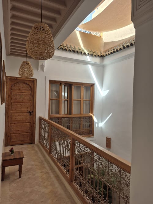 Couloir traditionnel du riad marocain avec luminaires suspendus tissés, porte en bois travaillé, balustrades sculptées et puits de lumière à Marrakech