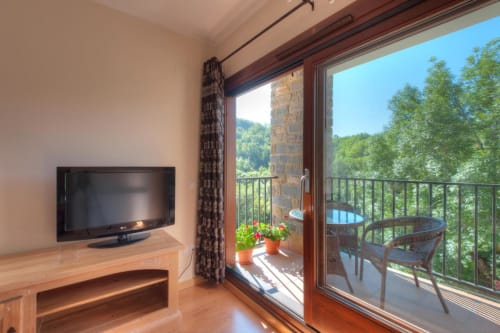 Chambre d'hôtel moderne avec TV écran plat, mobilier en bois et portes vitrées donnant sur balcon avec vue sur les montagnes vertes