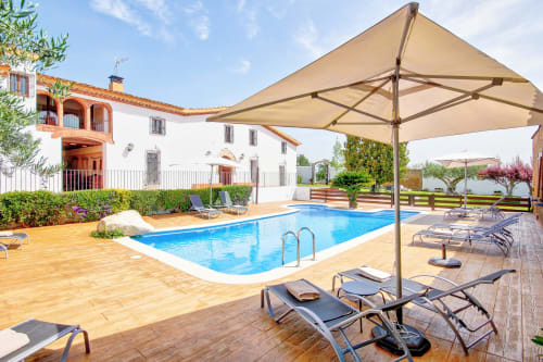 Área de piscina de la Masía Can Canyes con piscina rectangular azul, tumbonas, sombrillas beige y edificio rústico blanco