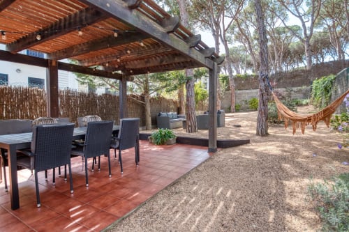 Shaded outdoor dining and lounge area in Palafrugell garden apartment with pergola, furniture, and tall trees