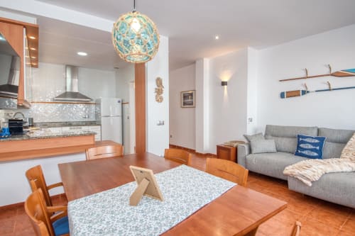 Modern apartment living area with turquoise pendant lamp, wooden dining table, gray sofa, and open kitchen with orange