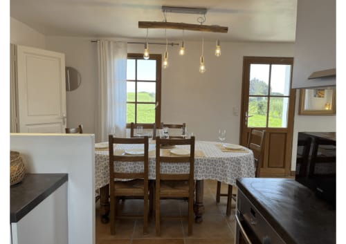 Salle à manger de cottage à Belle Île-en-Mer avec table en bois, luminaires suspendus rustiques et vue sur le jardin