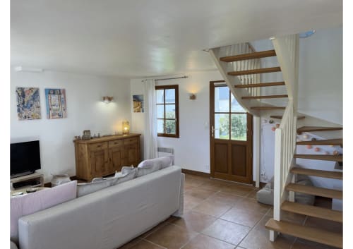 Salon lumineux avec canapé blanc, escalier en bois, téléviseur, commode et portes donnant sur le jardin à Locmaria Belle Île en Mer