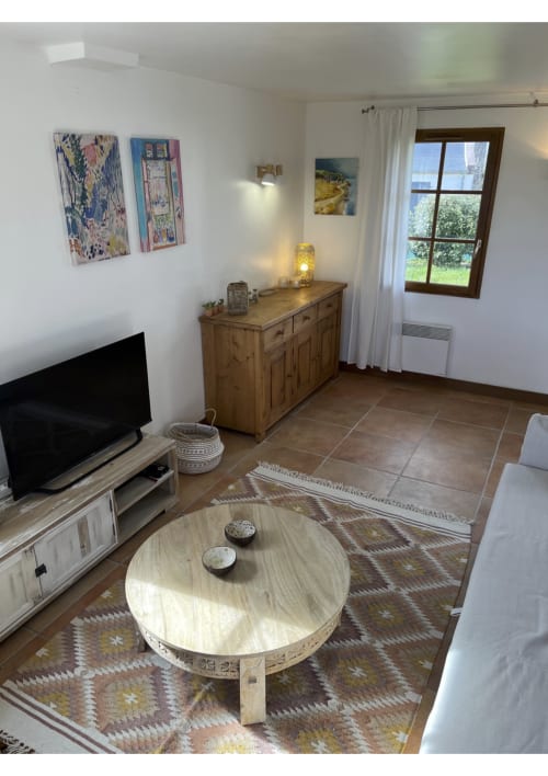 Lumineux séjour de chaumière avec mobilier en bois, téléviseur, table ronde, tapis à motifs et vue sur le jardin à Locmaria Belle Ile en
