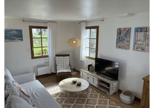 Lumineux séjour de chaumière avec vue sur le jardin, canapé blanc, télévision, table basse ronde, chaises en bois et lampe suspendue en osier