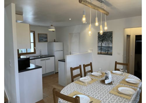 Salle à manger de cottage moderne avec suspensions en bois rustique, table dressée et cuisine ouverte à Loc'Maria Belle Île en Mer