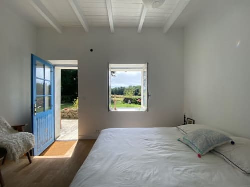 Chambre lumineuse avec vue sur le jardin par porte et fenêtre bleues, poutres blanches, parquet.