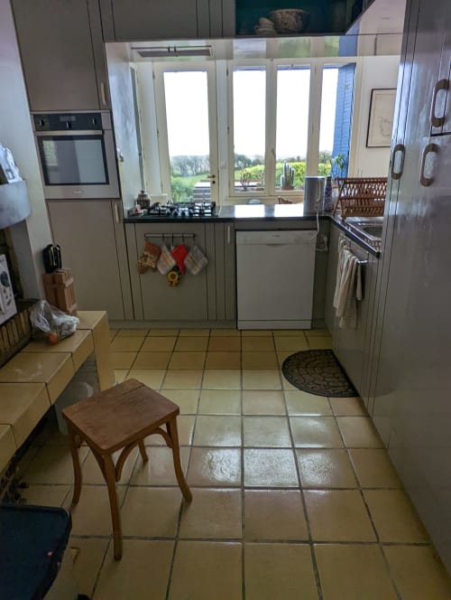 Cuisine de cottage avec portes vue mer, cuisinière à gaz, armoires blanches, carrelage et table en bois à Belle Île en Mer