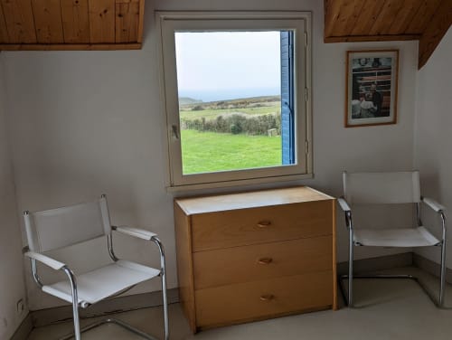 Chambre lumineuse de cottage avec plafond en bois, deux chaises blanches, commode en bois et grande fenêtre surplombant les champs verdoyants