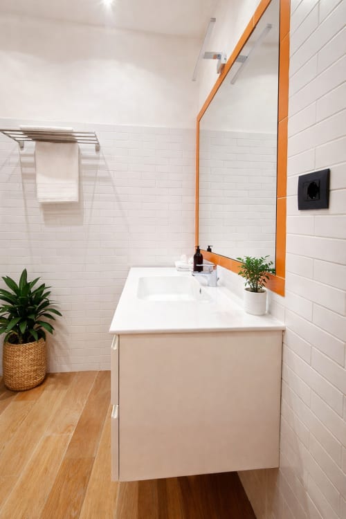 Bagno moderno a Zoelia Roma con piastrelle bianche, specchio con cornice arancione, mobile in legno, piante in vaso e portasciugamani