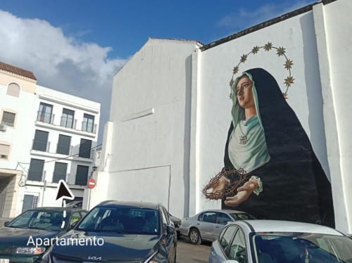 Gran mural de la Virgen María con aureola en la pared blanca de un edificio en Jerez de la Frontera, con autos estacionados abajo