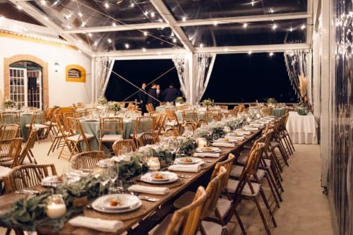 Sofisticada configuração de jantar ao ar livre em Quinta da Alcaidaria-Mór com cadeiras de madeira, mesas compridas, vegetação e iluminação de cordas