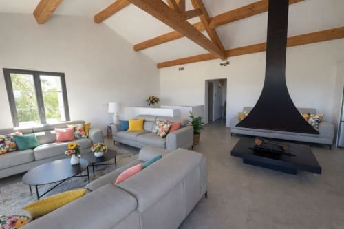 Villa moderne avec salon élégant, sièges gris, coussins colorés, poutres en bois apparent, cheminée noire et vue sur la mer