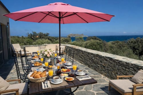 Terrasse bord de mer de la Villa Marine avec parasol rose, petit-déjeuner et vue panoramique sur la mer à Centuri, Corse