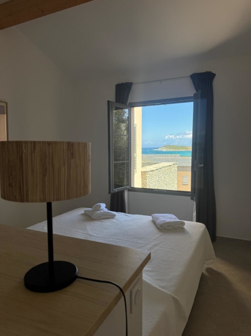 Chambre lumineuse de la villa La Conciergerie du Cap Corse avec vue mer sur le littoral corse par la fenêtre ouverte