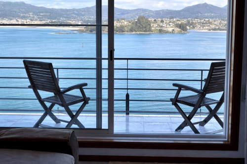 Dos sillas negras en una terraza con vistas al mar, la bahía de Baiona, montañas y pueblo costero de fondo