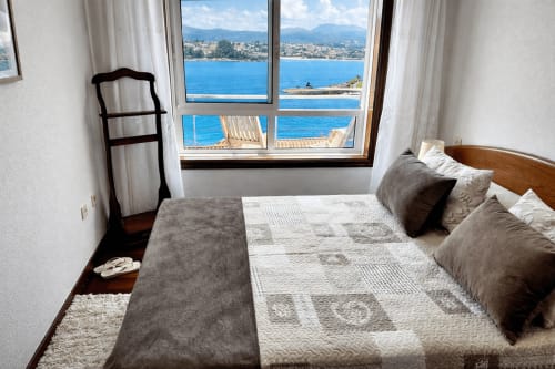 Dormitorio frente al mar en Holibai Baiona con vistas al mar, cama de madera y costa mediterránea visible a través de grandes ventanales