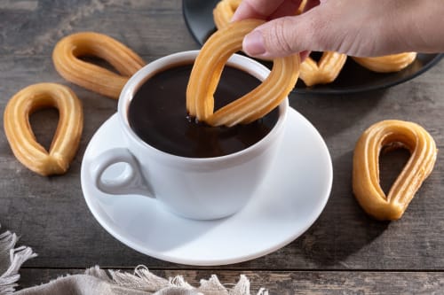 Mano sumergiendo un churro en salsa de chocolate en Ab Be Apartments, Málaga