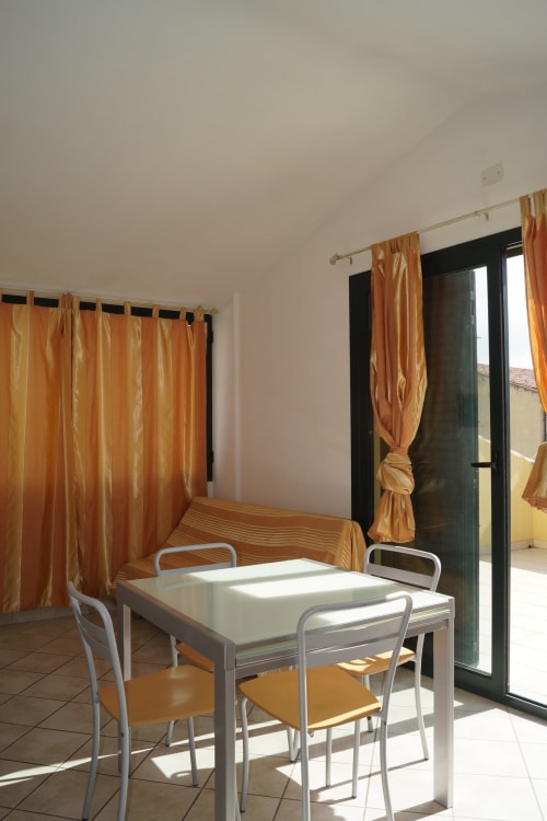 Bright apartment dining area in Orosei with white table, metal chairs, orange curtains, and glass doors opening to veranda