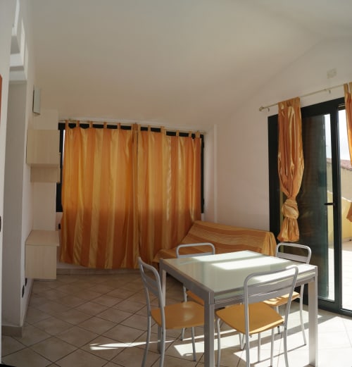 Bright apartment room in Orosei with orange curtains, white dining table, metal chairs, and glass door leading to veranda