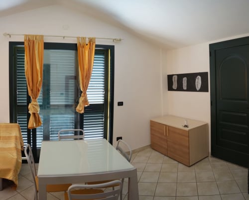 Modern apartment dining area with white table, wooden dresser, gold curtains, dark green doors and veranda access in Orosei