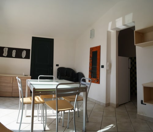 Bright apartment dining area with table, chairs, black door, orange-framed mirror, and white walls in Orosei, Italy