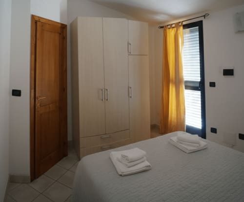 Bedroom in Orosei apartment with bed, large wardrobe, wooden door, and yellow curtains by window