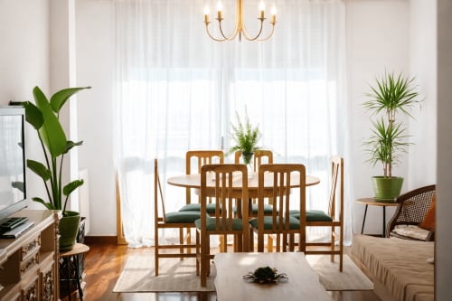 Comedor luminoso en el apartamento Amieiro de Baiona con mesa de madera, sillas, plantas de interior y vistas a la ciudad a través de cortinas traslúcidas