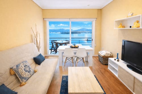 Luminoso salón amarillo con terraza vista al mar en apartamento de Baiona, mobiliario blanco y comedor con vistas a las montañas