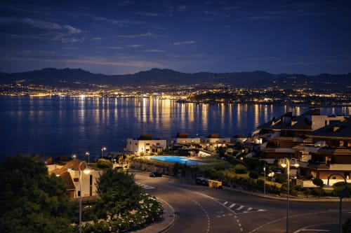 Baiona al atardecer con edificios iluminados, piscina y montañas reflejadas en aguas tranquilas