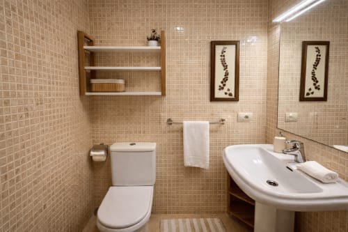 Baño moderno en apartamento A Son do Mar, Baiona. Inodoro, lavamanos pedestal, espejo, estantes de madera y paredes de azulejos beige.