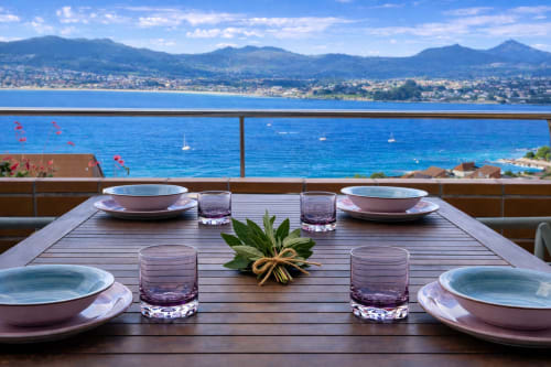 Mesa de comedor elegante en terraza con vistas al mar frente a la bahía de Baiona con veleros y montañas costeras, apartamento A Son do Mar