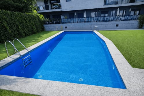 Piscina rectangular con azulejos azules en el apartamento Holibai de Baiona, rodeada de jardín cuidado y edificio moderno