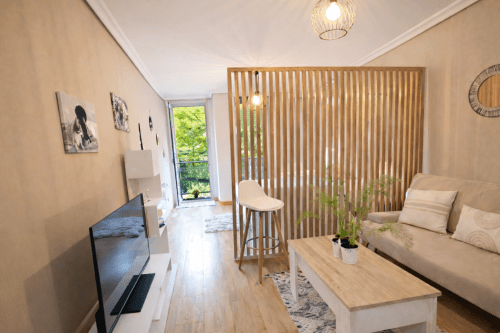Luminoso salón moderno en Baiona con sofá beige, divisor de madera, TV y acceso a balcón con vistas a la vegetación